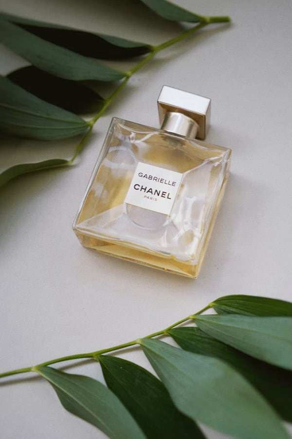 Flacon de parfum Gabrielle CHANEL posé sur une surface claire entouré de feuilles vertes – vue verticale