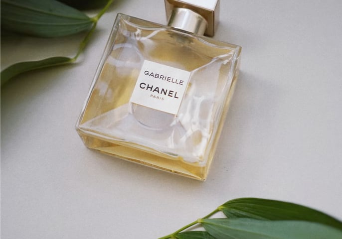 Flacon de parfum Gabrielle CHANEL incliné sur une surface claire avec des feuilles vertes – vue horizontale